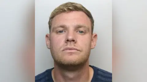 South Yorkshire Police Custody picture of Mason Lowe, He has short blonde hair and light-coloured stubble. He is wearing a nay t-shirt.