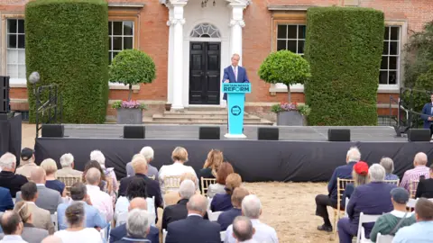 Qays Najm/BBC Nigel Farage addresses the crowds in front of a stately home, speaking from a Reform UK branded podium. There are neatly pruned bushes behind him, outside the stately home.
