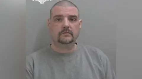 Chris Hemming pictured in a mugshot. He has a blank expression and wears a grey jumper. He has dark stubbly hair and a dark beard. 
