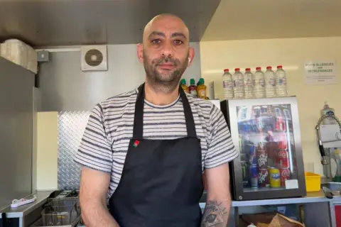 Ollie Conopo/BBC A man in a black and white striped T-shirt and a navy apron stands in a food truck. Behind him are lots of bottles of water and a transparent fridge with soft drinks in.