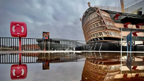 An image of the stern of the HMS Victory in its dock with a perfect reflection in water. A life ring in its housing is on the left of the shot, equally reflected. Railings and a building sit in the background.