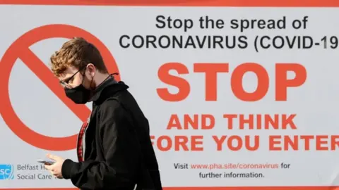 PA Media A young man wearing a mask looking at his phone and walking in front of a Covid-19 awareness poster from the Department of Health