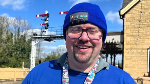 Emma Baugh/BBC James Rollo is wearing a blue hat which says Autism on it. He is also wearing a blue jumper and a Thomas the Tank Engine lanyard. He is standing in front of a small railway station.