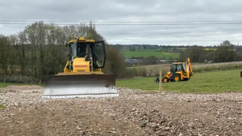 Two yellow construction vehicles dig grassland in a field. One bulldozer faces the camera