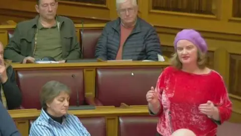 LDRS A woman in a red jumper and a purple beret is standing in a council chamber. The seats are burgundy and light wood. There are two men and a woman watching. 