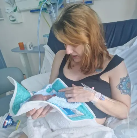Corrinne and Sam Boulton Corrinne Boulton who has shoulder length light brown hair and a fringe falling over eyes and looks distressed holds her tiny baby in a blanket - she is sitting up in a hospital bed. 