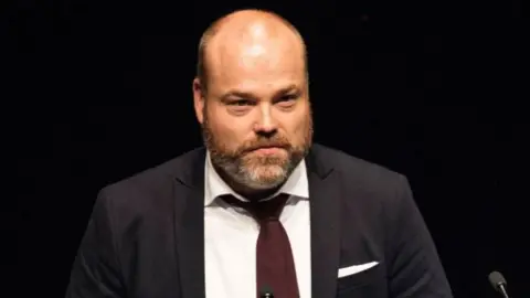 Getty Images a bald man with a brown beard wearing a dark suit, white shirt and purple tie