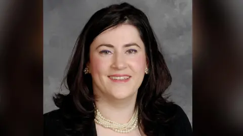 Sandra Stewart smiles at the camera. She has long brown hair and is wearing a pearl necklace and earrings and a dark coloured jacket.