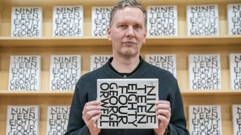 PA Media David Shrigley standing in front of a book shelf of his 1984 edition.