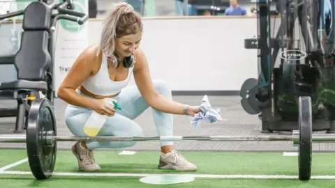 PA Media Laura Kendrew sanitises equipment during her workout at the PureGym in Leeds, Yorkshire, as indoor gyms, swimming pools and sports facilities can reopen as part of the latest easing of coronavirus lockdown measures in England.