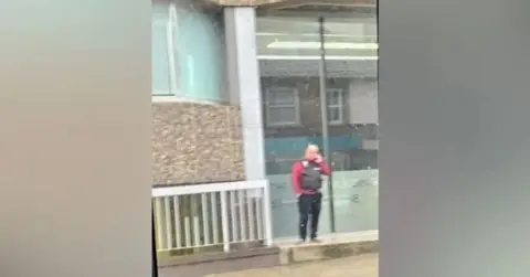 Submitted Blurry picture of a man wearing a tactical vest outside an office building. He is holding a mobile phone to his ear.