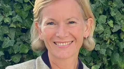 Hattie Lawrence A woman with short blonde hair wearing small earrings smiling at the camera in front of a bush