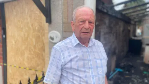Bob stands outside the burned remains of his home. He has a checked shirt on and short, grey hair.