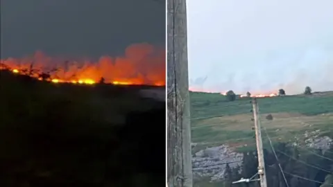 There are two images combined, on the left hand side is an image of a wildfire burning on a grassy mountain at night. The image on the right is the same mountain burning during the day time, there is also a large telephone pole in the foreground of the left image. 