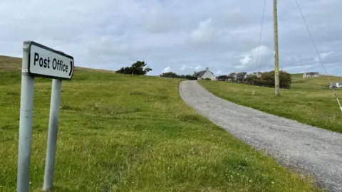 A road sign pointing to the Post Office. It is mounted above a grass verge, next to a narrow road. A house and trees are visible on the horizon.