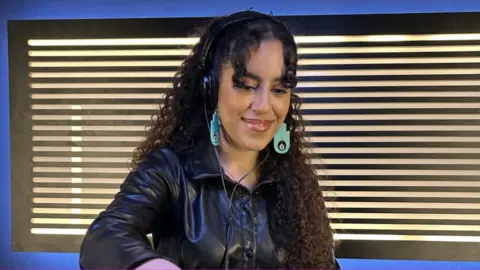 BBC Nooriyah smiles as she mixes in a recording studio. She's got shoulder-length brown curly hair, with large, dangly cyan-coloured earrings in a Hand of Fatima design. That's a flat with an eye in the middle of the palm. Nooriyah's also wearing over-ear headphones and a black leather jacket.