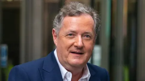 PA Media Broadcaster Piers Morgan. His head and shoulders are seen as he walks in front of a set of revolving doors. He is wearing a blue suit.