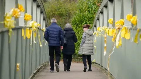 PA Media Nicola Bulley's sister, Louise Cunnigham (right), with her mother and father-in-law walking past the yellow ribbons and messages of hope tied to a bridge over the River Wyre