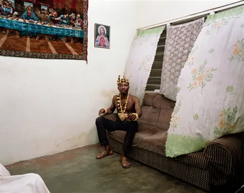 Deana Lawson A photo by Deana Lawson of a shirtless man sitting on a sofa, with religious images beside him on the wall