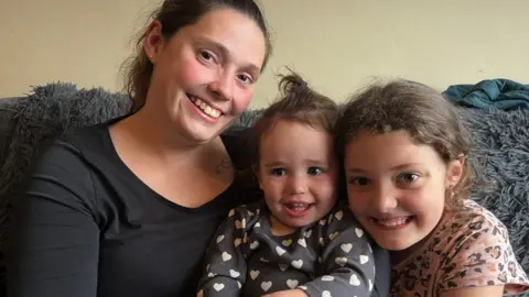 Gina Baker with two of her three children. She has her arms around the youngest, who is sat in the middle. They all have dark hair and dark eyes. They are sat close together on the sofa with a grey blanket behind them. They are all smiling and looking at the camera.