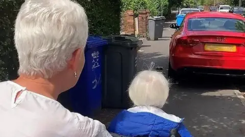Woman pushing a woman using a wheelchair on the pavement blocked by a parked car