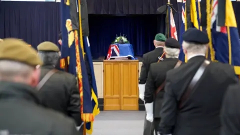 John Fairhall/BBC The aisle of the crematorium with the coffin draped in a flag. On either side of the crematorium veterans and serving soldiers stand.