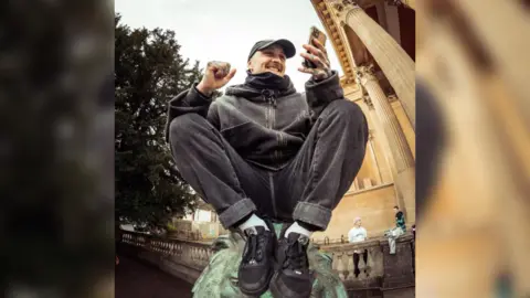 Ivaylo Erev  Danny Pierce sitting on top of a fountain statue and smiling at his phone