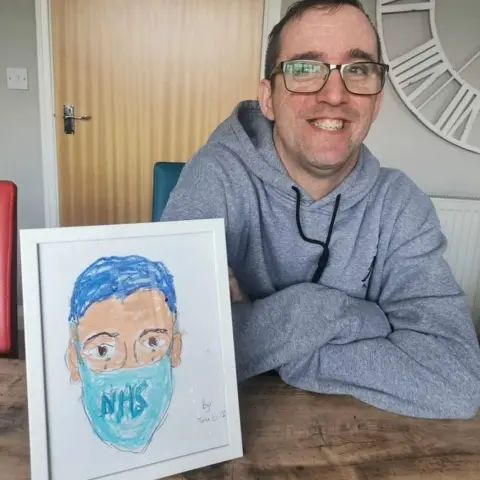 Tom Wilson Tom at a table with a frame of his portrait of an NHS worker wearing a mask