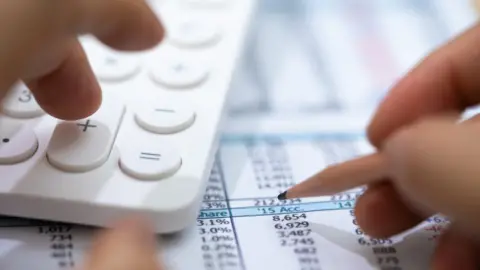 Getty Images A stock photo of a left hand using a calculator and a right hand holding a pencil hovering over a spreadsheet