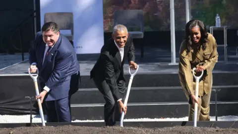 Illinois governor JB Pritzker digging dirt at the site in a ceremonial groundbreaking ceremony with Barack and Michelle Obama.