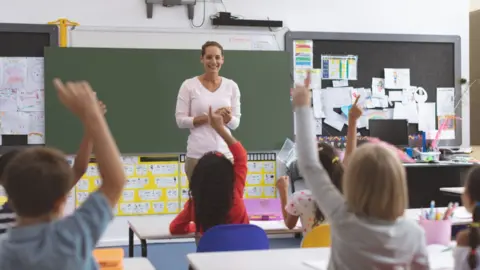 Getty Images Rear view of school kids raising hand while they are sitting on their chair with their teacher in front of them against a greenboard in background