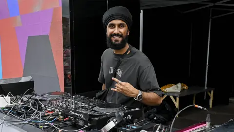 Virdi, an Asian man wearing a black turban representing his Sikh faith. wearing a grey tshirt. He's on DJ decks with multiple wires, buttons and faders.