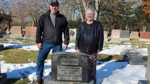 Dunn family Man and woman stand next to grave stone as snow melts on ground