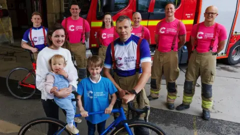 Cancer Research UK Ms Parsons with her husband Matt, their two young children and six firefighters from Avon Fire and Rescue.