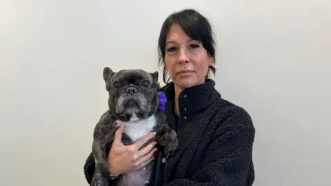 Robert Constantin/BBC Black and white French bulldog wearing purple bow and being held by woman with medium-length black hair