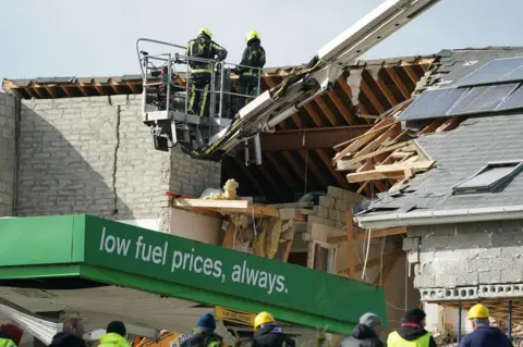 PA Media Firefighters on a mechanical platform assess the damage caused to the service station and a nearby apartment block