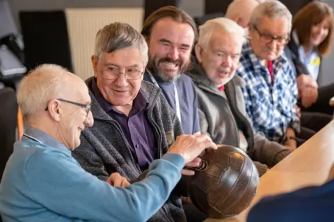 Sporting Memories Foundation An elderly man smiles as he picks up an old football as part of a Sporting Memories session