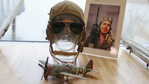 A photo of a pilot on display with a model plane, flight hat and glasses