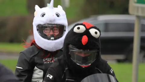 Andy Simons Rider with character helmet showing a black face with white eyes and a red nose, pillion passenger with white helmet with two small eyes