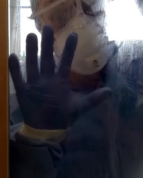  Sara Danesin An NHS worker in PPE equipment holds their hand up to the glass in a door