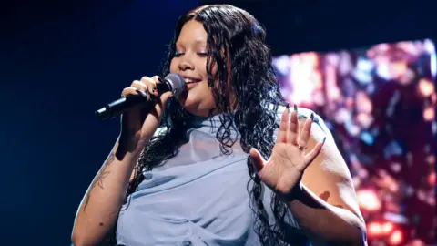 Getty Images Shygirl performing at the Mercury Prize. Real name Blane Muise, Blane wears a pale blue chiffon dress which drapes around her neck. She has long, dark wavy hair which she wears loose over her shoulders. She holds a microphone to her face with her right hand, revealing a rose forearm tattoo, and holds her left palm up. Dark staging can be seen behind her and she is lit by a spotlight.