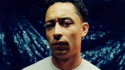Sirus Gahan Loyle Carner standing looking at the camera wearing a white T-shirt