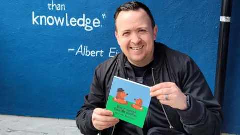 Luke Newman Luke Newman holding a book that he has written called the mental health moles