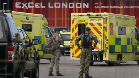 Getty Images Military personnel stand near London Ambulance Service vehicles at the new NHS Nightingale Hospital at ExCeL London on March 25, 2020 in London, England.