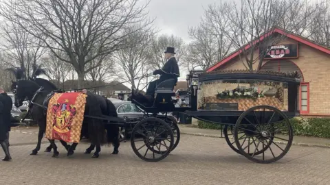 A horse drawn hearse leaves carrying the body of Leah Croucher. The horses are draped in a Harry Potter, Gryffindor banner with Leah's name on it.