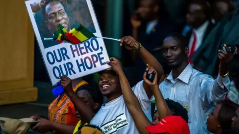 Getty Images Supporters hold banner, wave Zimbabwean national flag and cheer as they gather to welcome Zimbabwe's incoming President Emmerson Mnangagwa upon his arrival at Zimbabwe's ruling Zanu-PF party headquarters in Harare on November 22, 2017