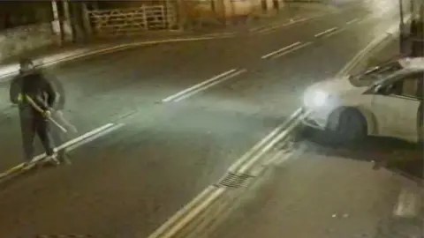 CCTV footage of one suspect standing in the road outside the shop with a baseball bat and the white Ford ST used by the suspects