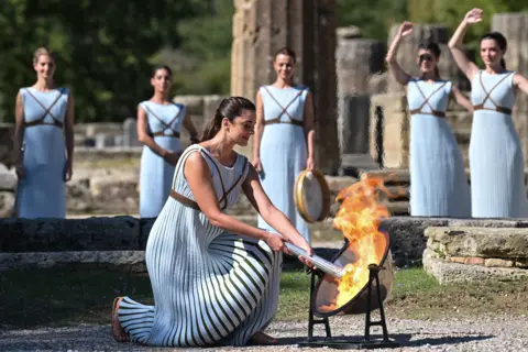Aris Messinis / AFP Greek actress Xanthi Georgiou, playing the role of the High Priestess, lights up the torch during the flame lighting ceremony for the Beijing 2022 Winter Olympics at the Ancient Olympia archaeological site, on 18 October 2021