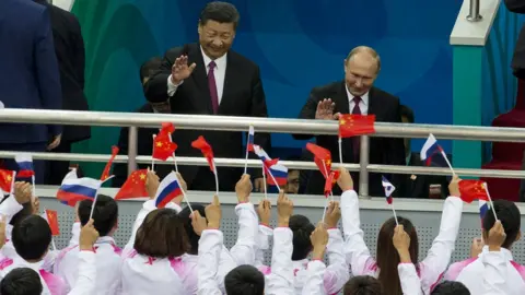 Getty Images Russian President Vladimir Putin (R) and Chinese President Xi Jinping wave to spectators during a friendly match between Chinese and Russian youth Ice Hockey teams on June 8, 2018 in Tianjin, China