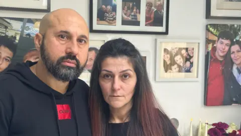 Jamie Niblock/BBC George and Areti Nicolaou in front of a wall of family pictures